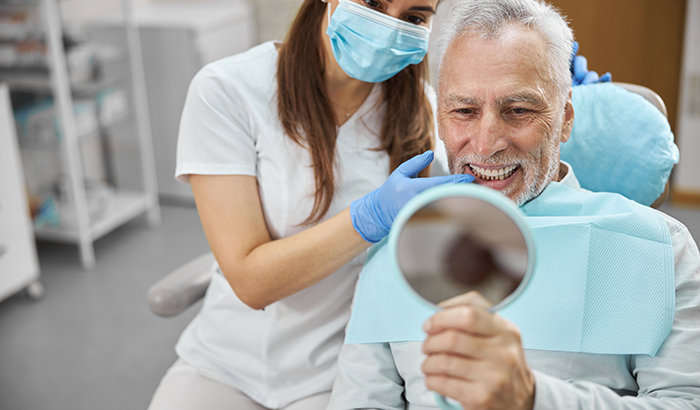Man holding mirror to his teeth and smiling in dental chair after receiving dental implants from Lanier Valley Dentistry in Dacula, GA